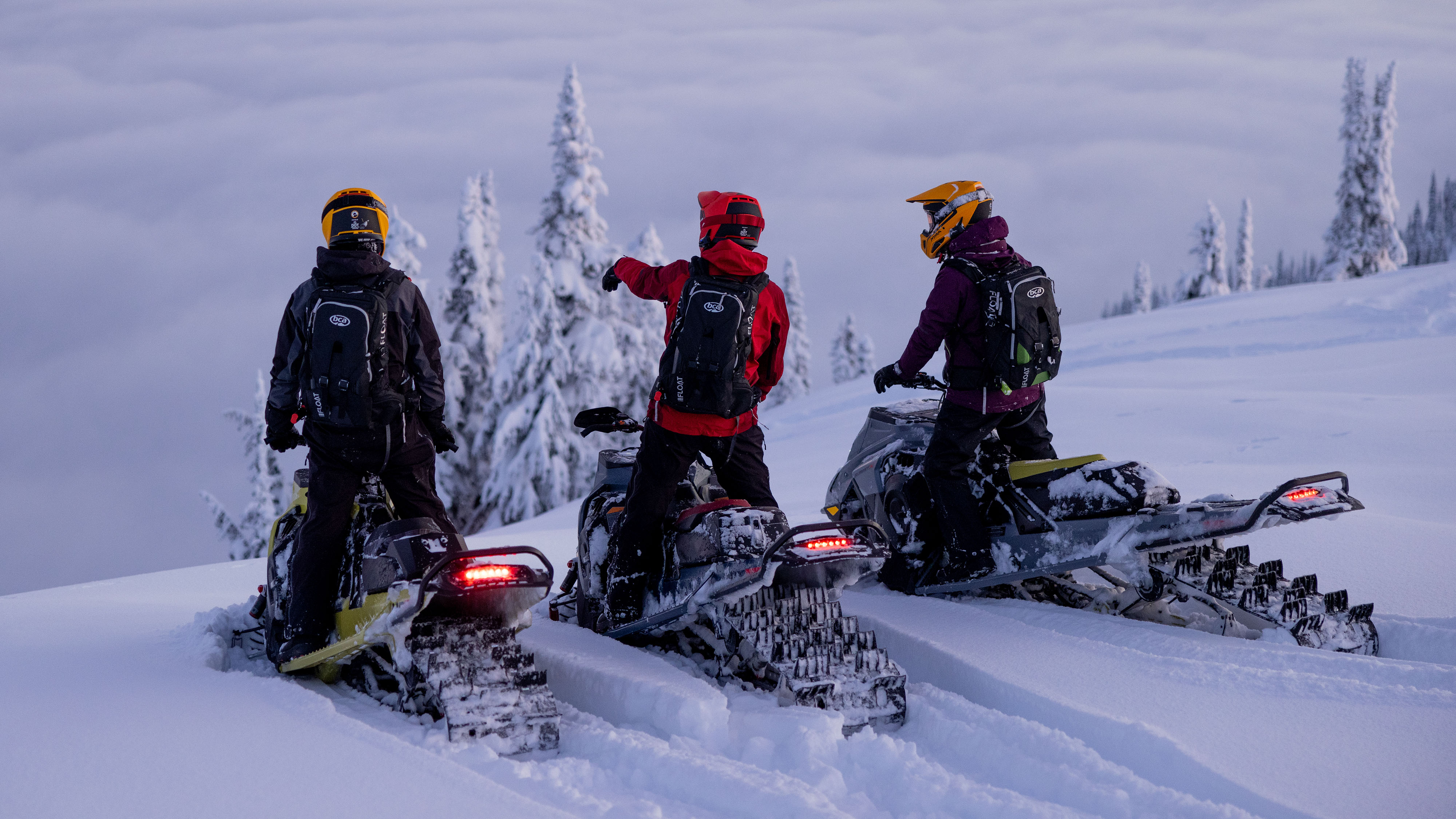 Kolme Ski-Doo-kuljettajaa seisoo moottorikelkkojensa päällä.