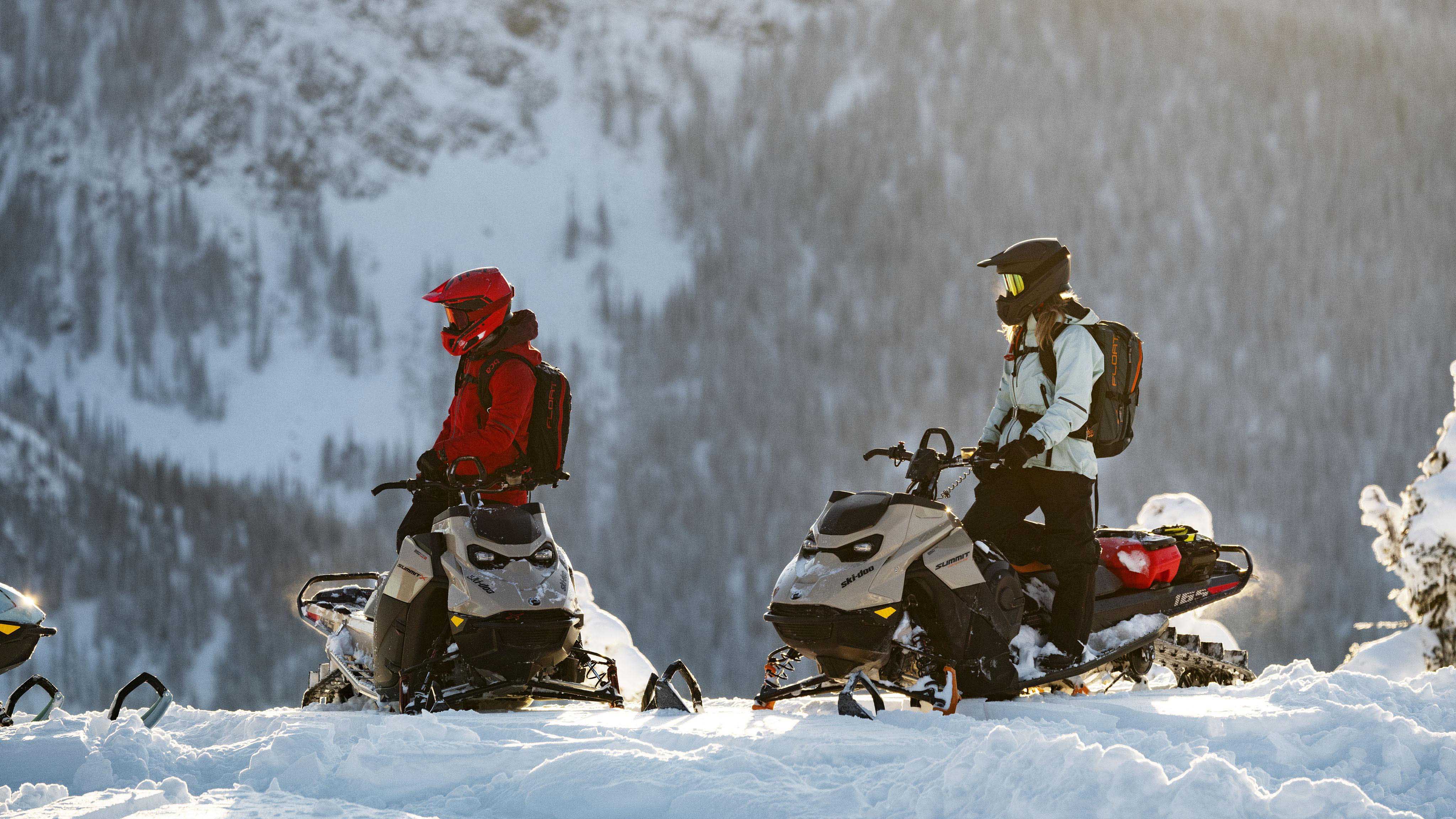 Two women standing on their Ski-Doo sleds at the top of a mountain