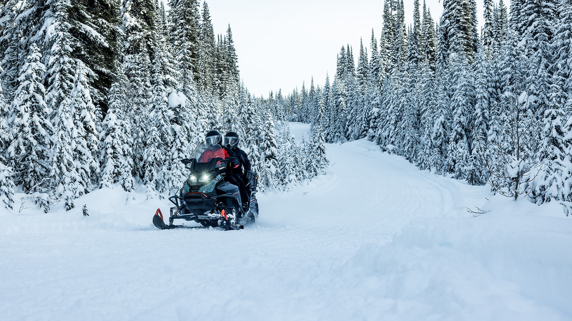 Two people on a 2026 Ski-Doo snowmobile riding through a snowy forest.