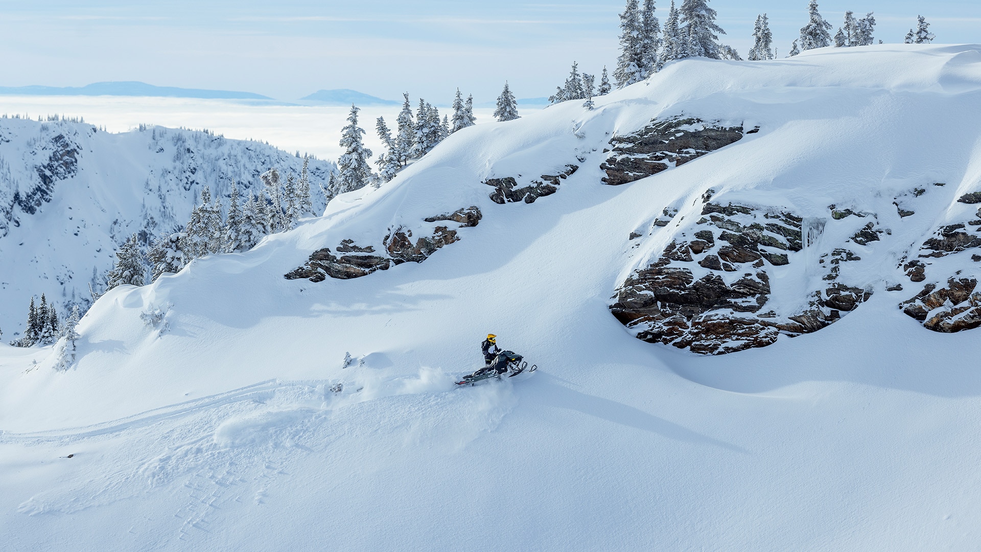 Un motoneigiste sur sa motoneige Ski-Doo Summit 2026 remonte une pente de montagne enneigée avec des rochers et des arbres éparpillés.