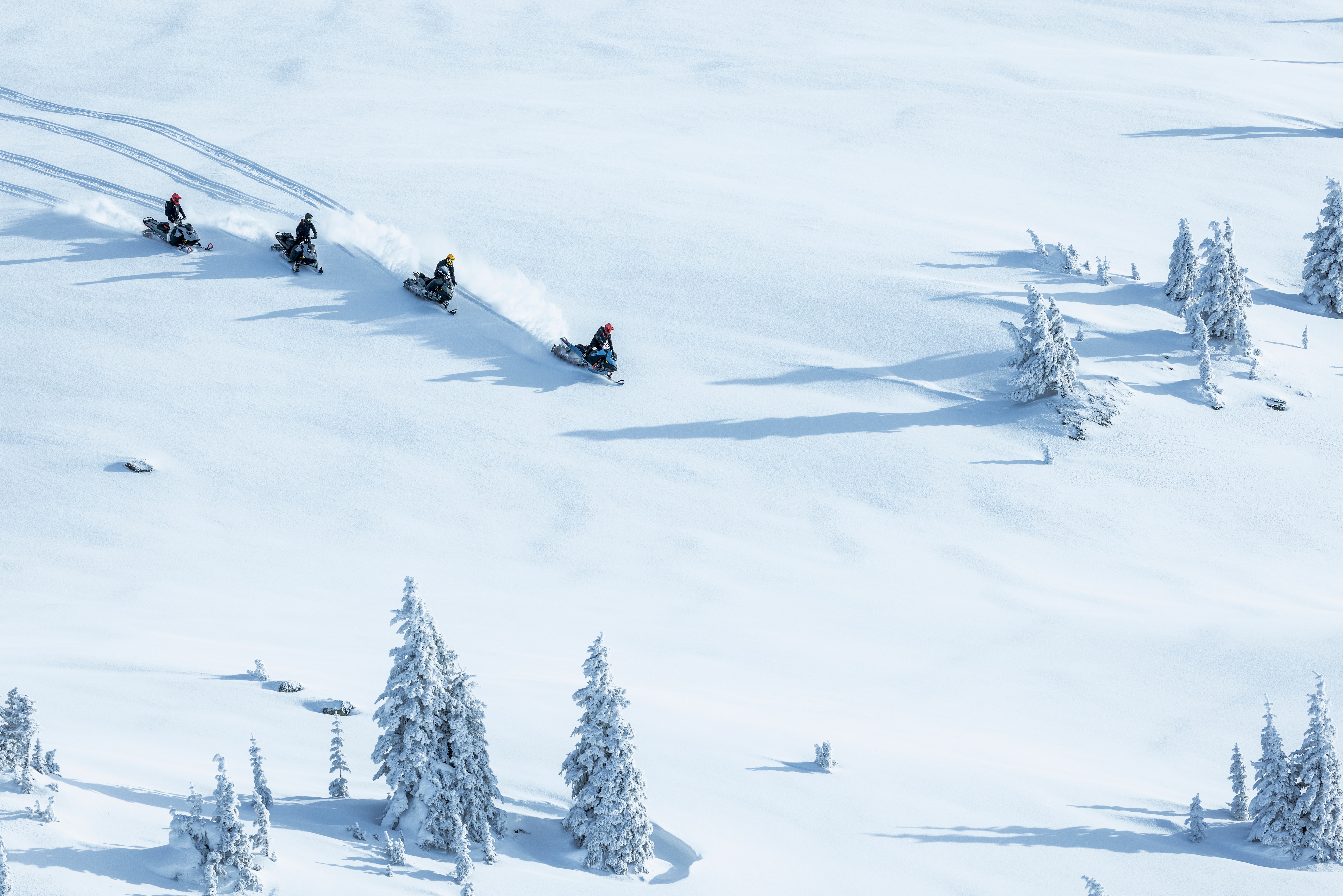 Four Ski-Doo snowmobiles riding across an open snowy field, leaving tracks in the fresh powder.