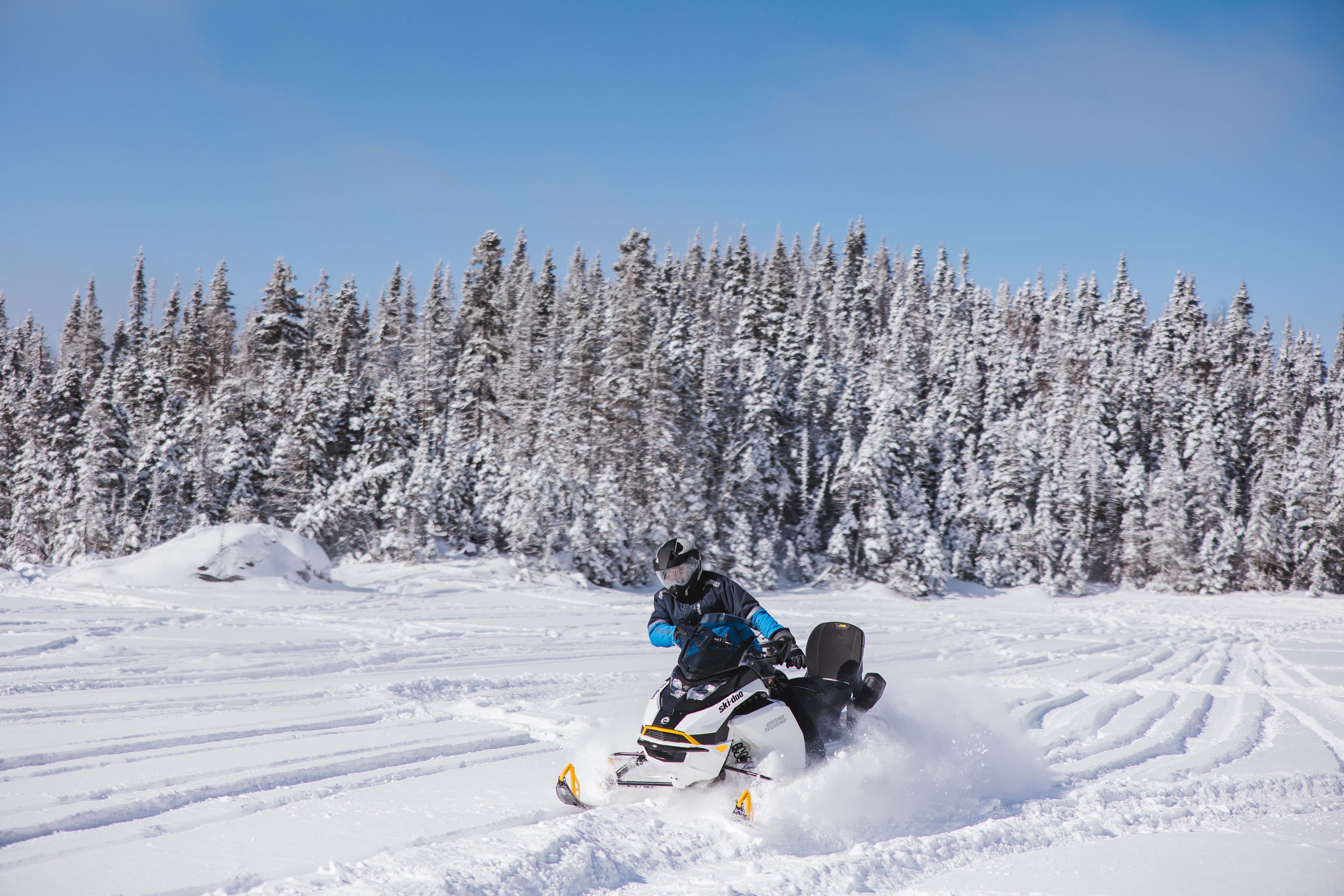 Snowmobiler riding a Ski-Doo Grand Touring snowmobile carving through fresh snow on a trail surrounded by snow-covered trees.