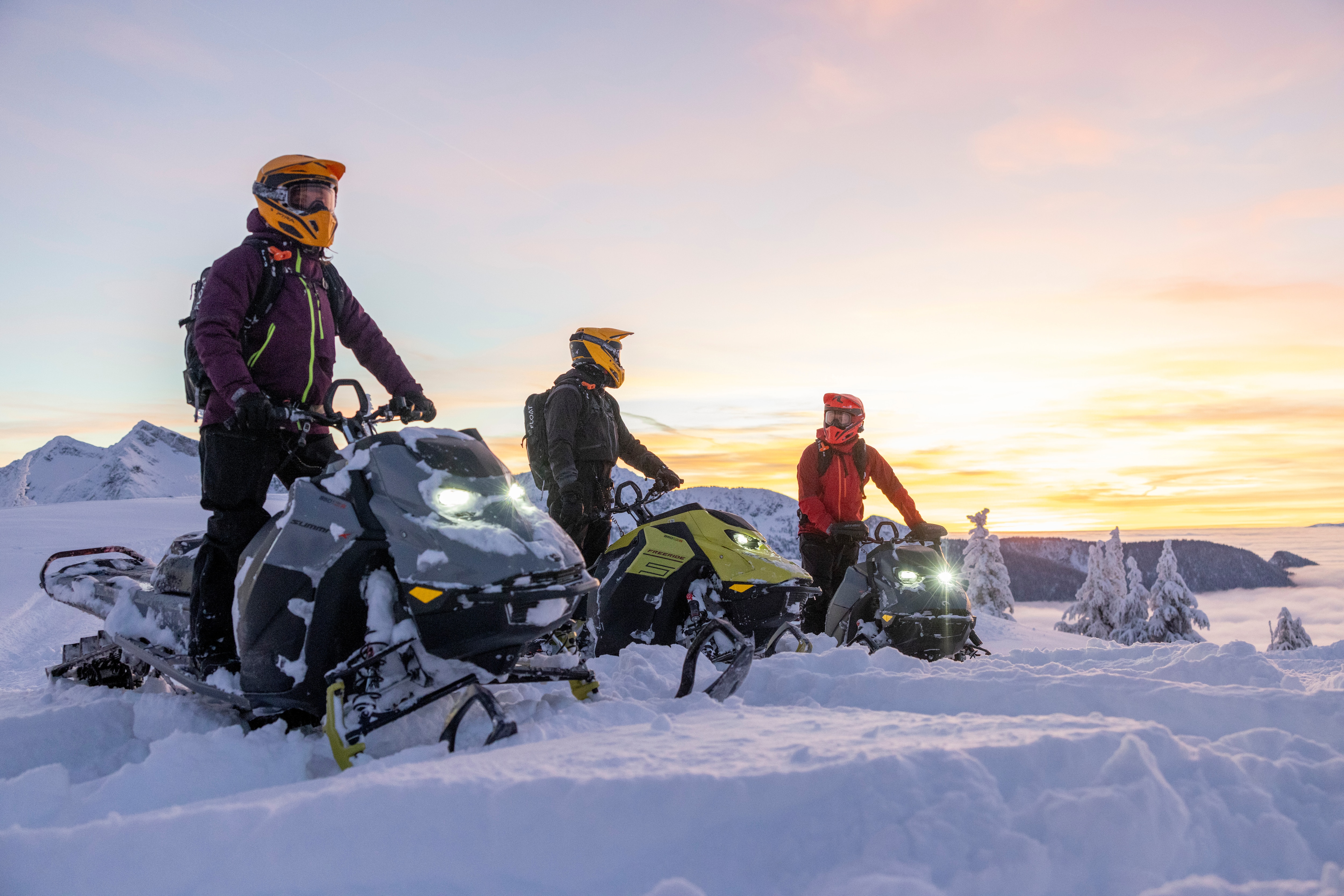 Three riders at the top of a mountain on their Ski-Doo snowmobile.