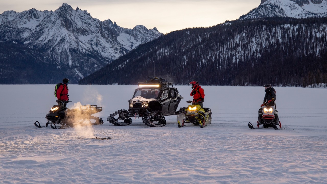 Quatre personnes en motoneiges Lynx et un véhicule tout‑terrain se rassemblent sur un paysage enneigé et gelé au crépuscule, entourés de montagnes alpines, pour une aventure hivernale en plein air.